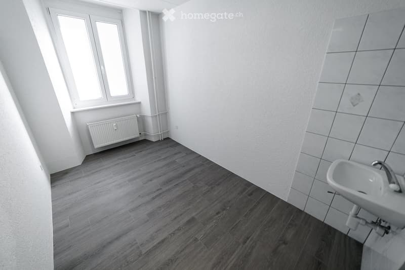 Empty room with wooden flooring, white walls, a radiator, and a window with white frames. A small sink is visible in the corner.