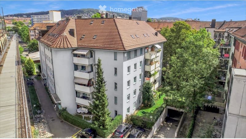 Multi-story residential building with a red-tiled roof, balconies, and surrounding greenery. Includes a parking area and garage.