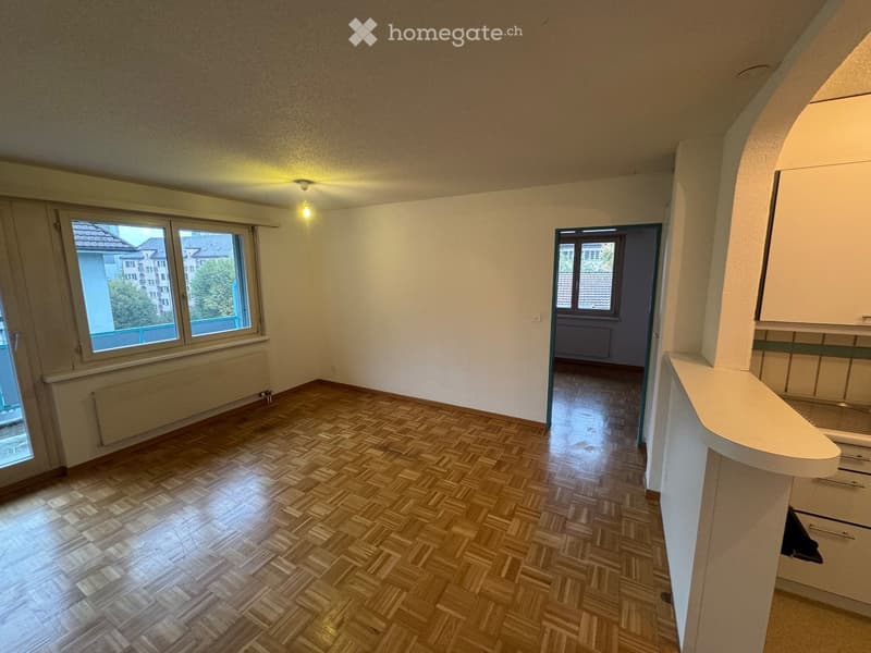 Empty room with wooden herringbone flooring, white walls, and large windows offering a view of neighboring buildings. The room features a built-in kitchen counter on one side and a doorway leading to another space.
