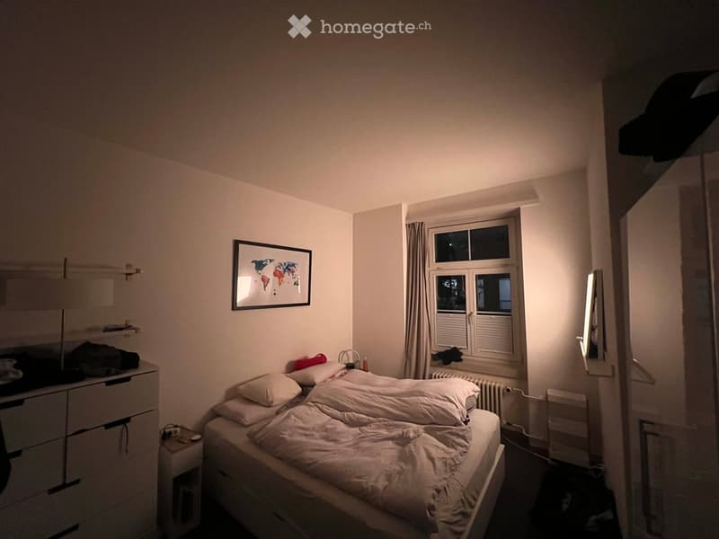 A small, neatly arranged bedroom with a single bed, pillows, a nightstand, a drawer unit, a picture frame on the wall, and a window with curtains.