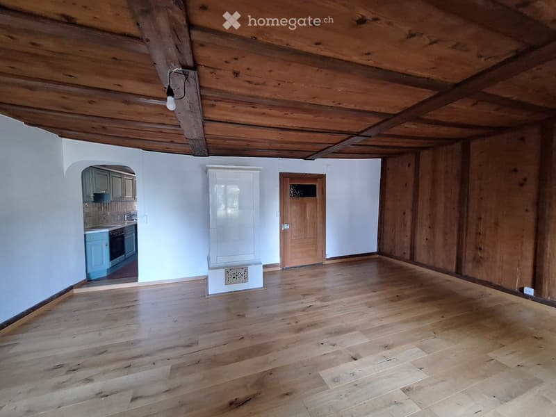 Empty room with wooden floor and exposed wooden beams on the ceiling. There is a doorway leading to another room with cabinets and appliances visible.