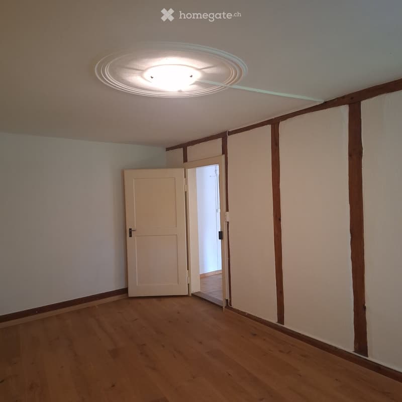 Empty room with wooden flooring, white walls, and a ceiling light fixture. A door leads to another room.