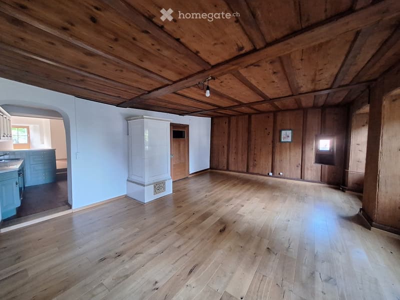 An empty attic space with wooden beams on the ceiling, wooden walls, and a polished wooden floor. The room has exposed wooden beams and a single light fixture hanging from the ceiling. There is a doorway leading to another room with blue cabinets and a si