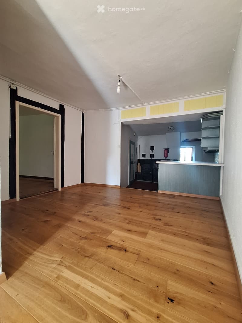 Empty room with wooden flooring, white walls, and a doorway leading to another room. The adjacent room has a kitchen area with cabinets and a countertop.
