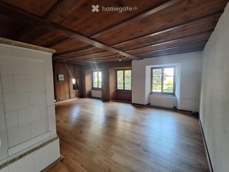 Empty room with wooden floors, exposed wooden ceiling beams, and large windows allowing natural light. The walls are painted white, and there are radiators beneath the windows.