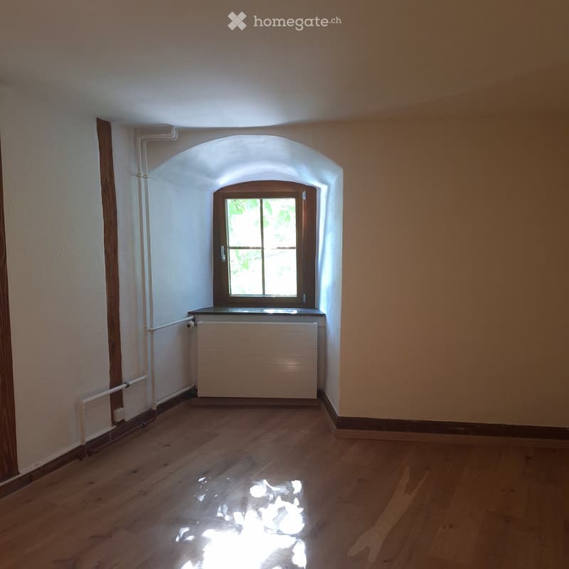 Empty room with wooden flooring, white walls, and a single window with wooden frame. Radiator is visible below the window.