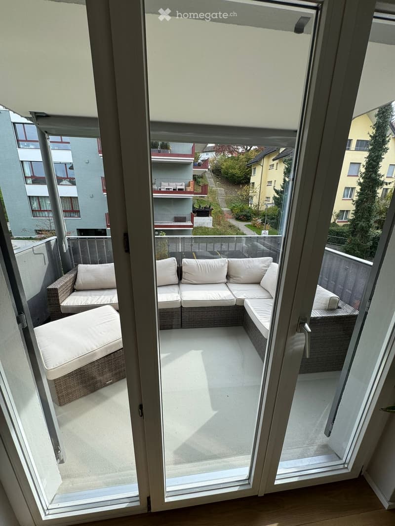Balcony with a modern, light-colored sectional sofa and a matching chair, featuring a glass railing and a view of neighboring buildings and greenery.