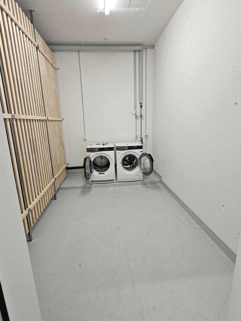 Minimalist laundry room with white washing machine and dryer against a concrete wall, wooden paneling on one side, and a concrete floor.