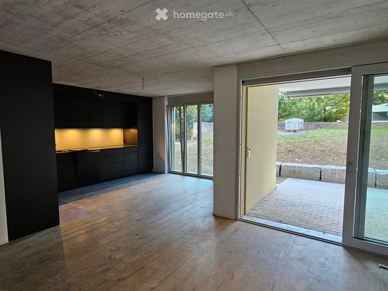 An unfurnished room with wooden flooring, large sliding glass doors leading to an outdoor area, and a dark kitchen area visible in the background.