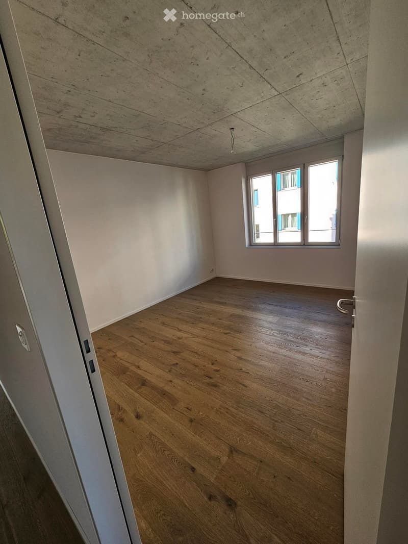 Empty room with wooden flooring, white walls, and a large window letting in natural light. The ceiling has a unique, slanted design.