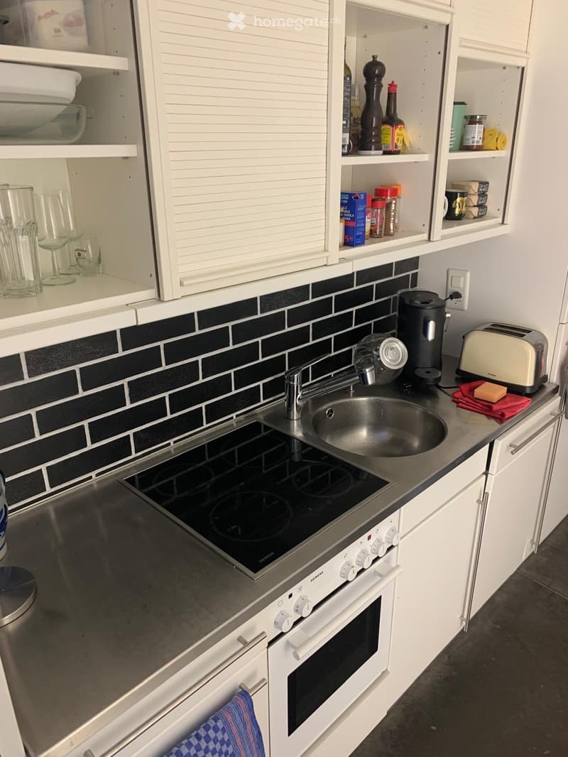 Modern kitchen with white cabinets, stainless steel sink, induction cooktop, oven, coffee maker, and toaster. Shelves hold various kitchen items including bottles, cups, and food containers.