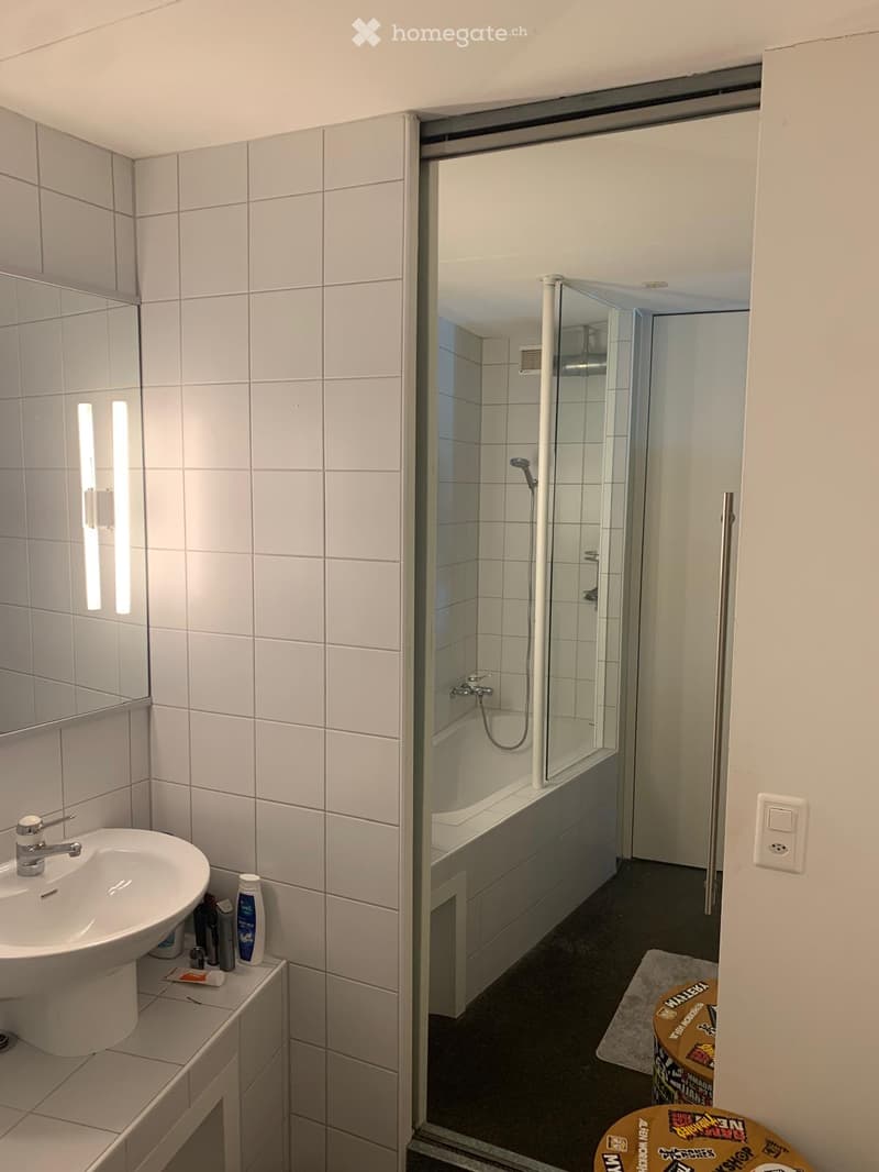 Modern bathroom with white tiled walls, a sink with a modern faucet, a bathtub with a glass shower enclosure, and toiletries on the sink counter.