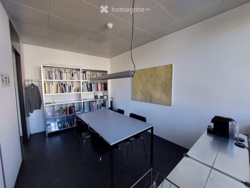 Modern office space with a long conference table, chairs, bookshelf filled with books, a coat hanging on a rack, and a printer on a side table.
