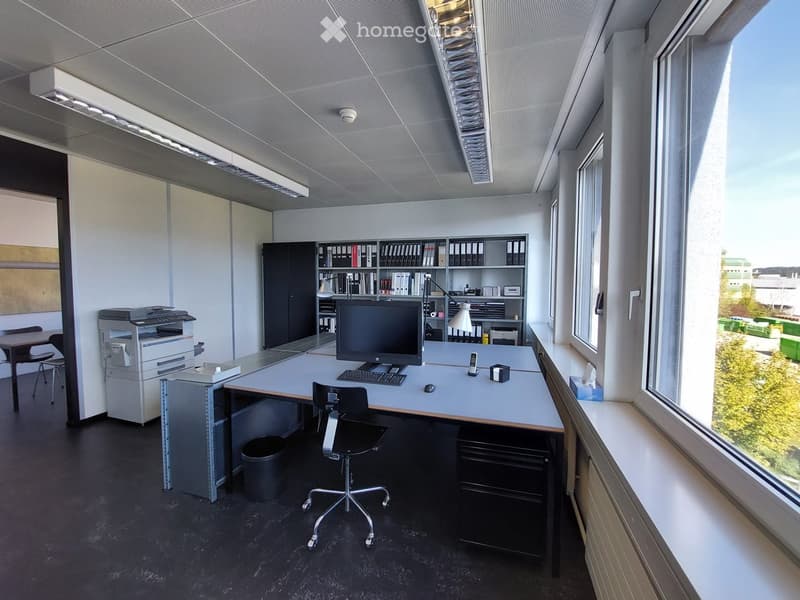 Modern office space with a desk, chair, computer, keyboard, mouse, printer, cabinet, bookshelf, and lamp. Large windows provide natural light.
