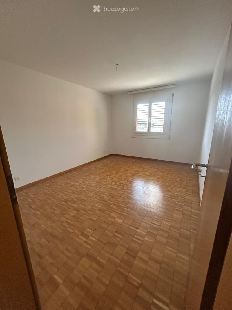 Empty room with wooden flooring and white walls, featuring a single window with blinds and a door frame visible on the left side.