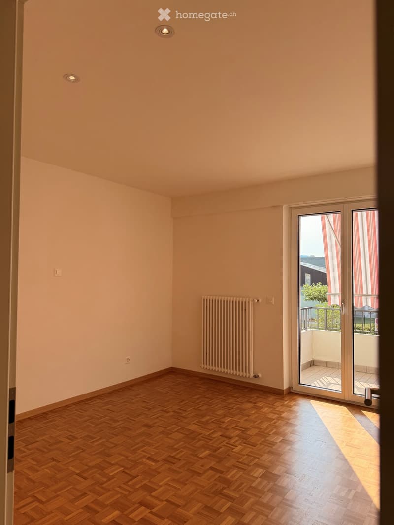 Empty room with wooden parquet flooring, light-colored walls, and a radiator. A sliding glass door leads to a balcony with outdoor furniture visible.