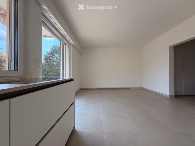 Empty room with white walls, tiled floor, and large windows offering a view of trees. There is a built-in cabinet along one wall.
