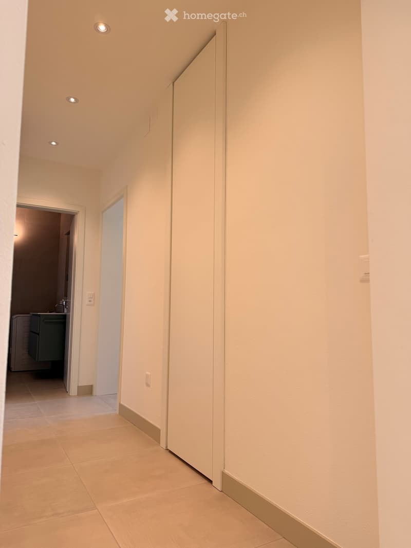 Empty hallway with beige tiled flooring, white walls, and recessed ceiling lights. Features a closed white door on the right and an open doorway leading to another room on the left.