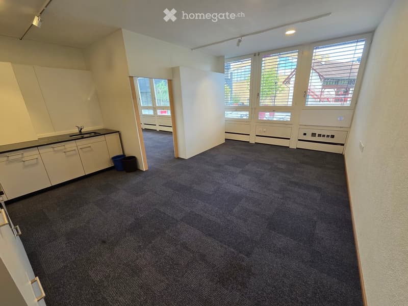 A small, furnished room with a sink, cabinets, and trash bins. The room has carpeted flooring and large windows with blinds.