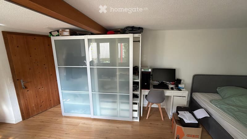 A furnished bedroom with a bed, desk, chair, computer, wardrobe, and a wooden door.