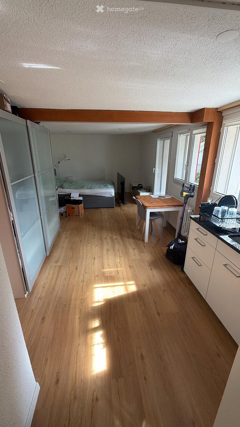 A well-lit room with wooden flooring, a small kitchen area, a bed, a table with chairs, and a refrigerator. The space is organized and features natural light from windows.
