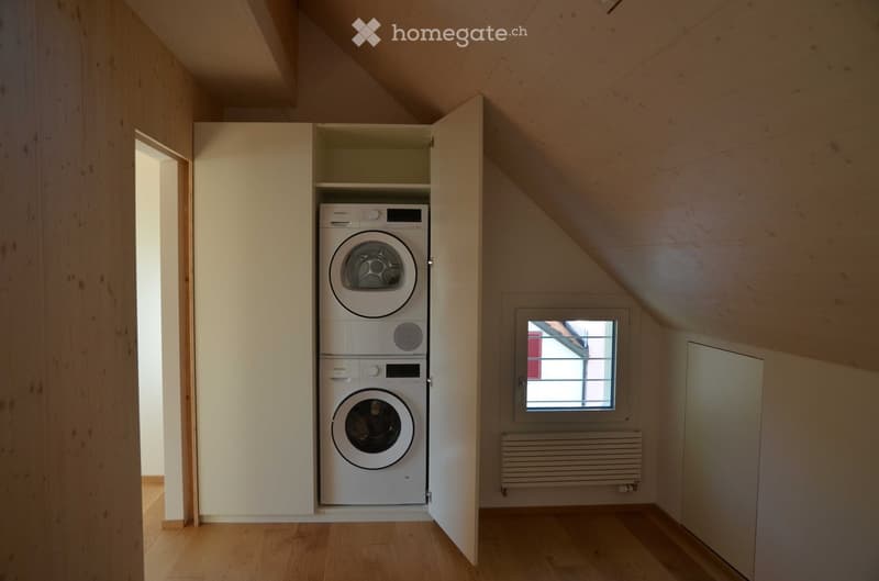 Attic room with wooden flooring, white walls, and a built-in cabinet housing a stacked washing machine and dryer. A small window with a view of a neighboring building is visible.