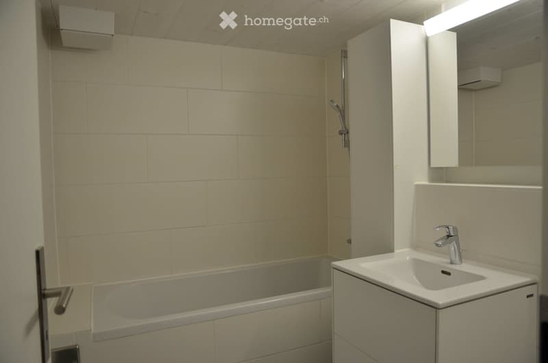 Modern bathroom with a bathtub, sink, mirror, and cabinet. The walls are tiled in a light beige color, and the fixtures are sleek and contemporary.