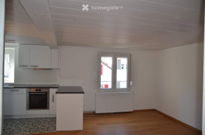 Empty kitchen with white cabinets, a black countertop, and a built-in oven. The room features wooden flooring and large windows with a view of the outside.