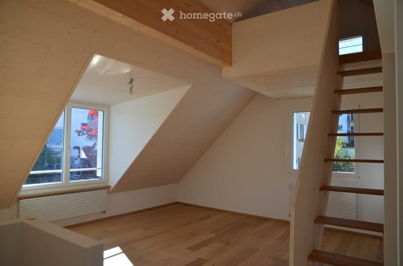 Bright, empty attic room with wooden floors, white walls, and exposed wooden beams. Features a staircase leading upwards, large windows with views, and built-in radiators.