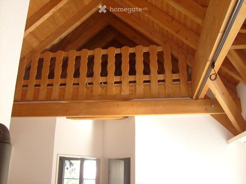 View of a wooden loft area with exposed wooden beams and a wooden railing, white walls, and a window showing outdoor view.