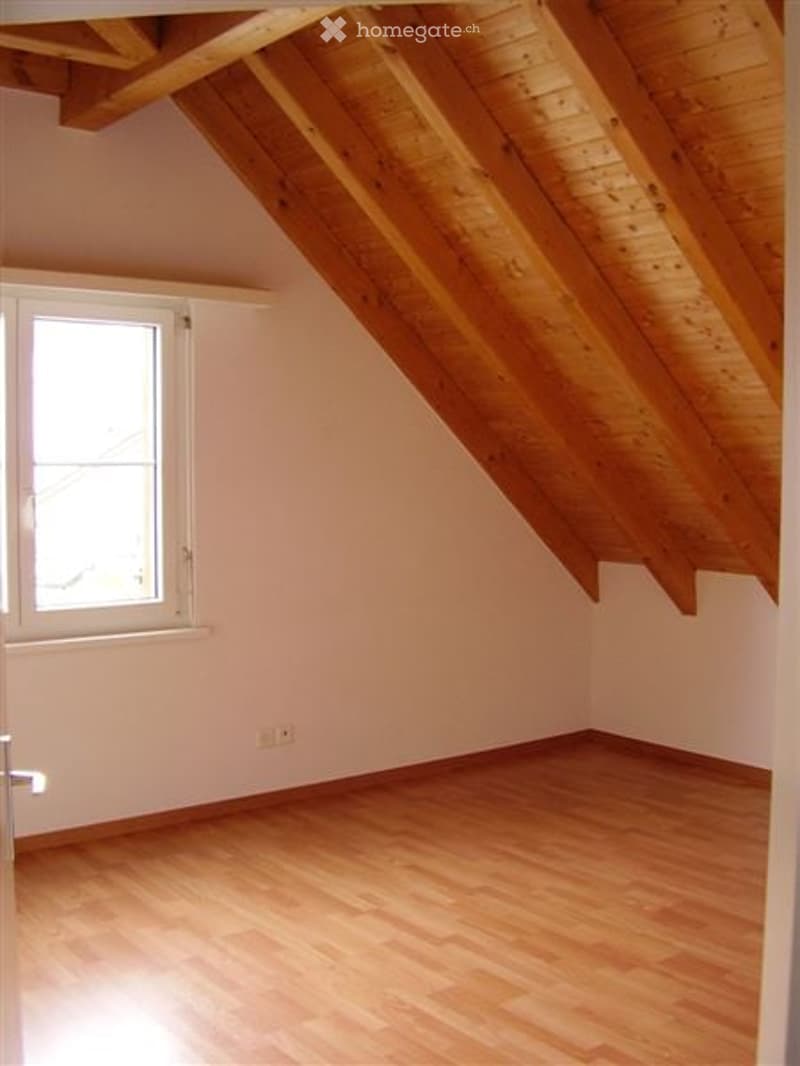 Empty attic room with wooden slanted ceilings and light wooden flooring, featuring a single window and white walls.