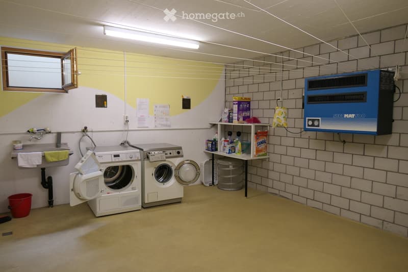 A small laundry room with a washing machine, dryer, sink, and various cleaning supplies on a shelf. The room has a window, tiled walls, and a small air conditioning unit.