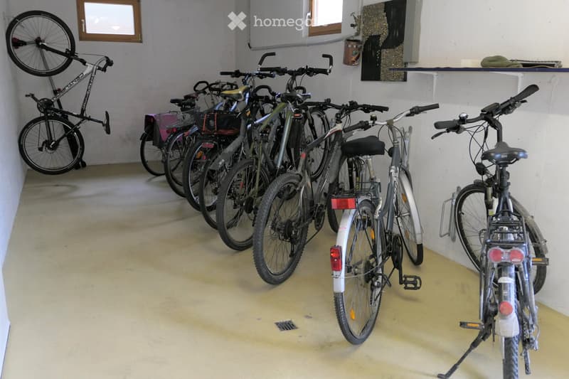 Empty room with concrete floor and white walls, several bicycles stored inside, one mounted on the wall, others standing on the floor.