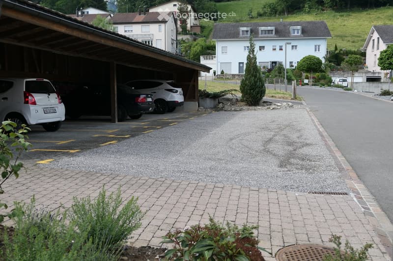 A covered garage with several parked cars, surrounded by a paved area and a residential street with houses in the background.