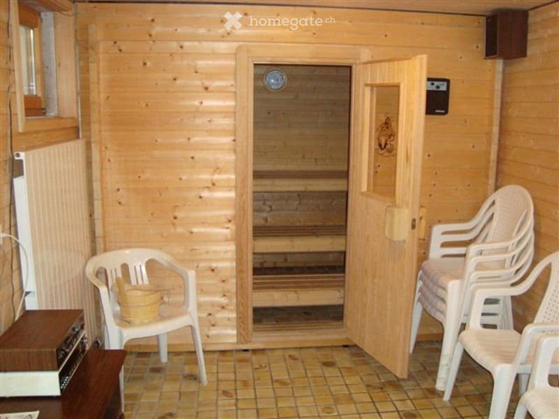 A wooden-walled room with a sauna door open, showing wooden benches inside. There are several white plastic chairs and a small table with a basket on it.