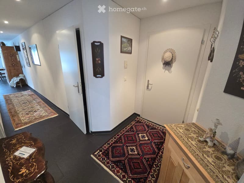 A well-lit hallway with white walls, dark tiled flooring, and several area rugs. There are framed pictures on the walls, a small wooden table with a decorative item, and a coat rack with a hat hanging on it.