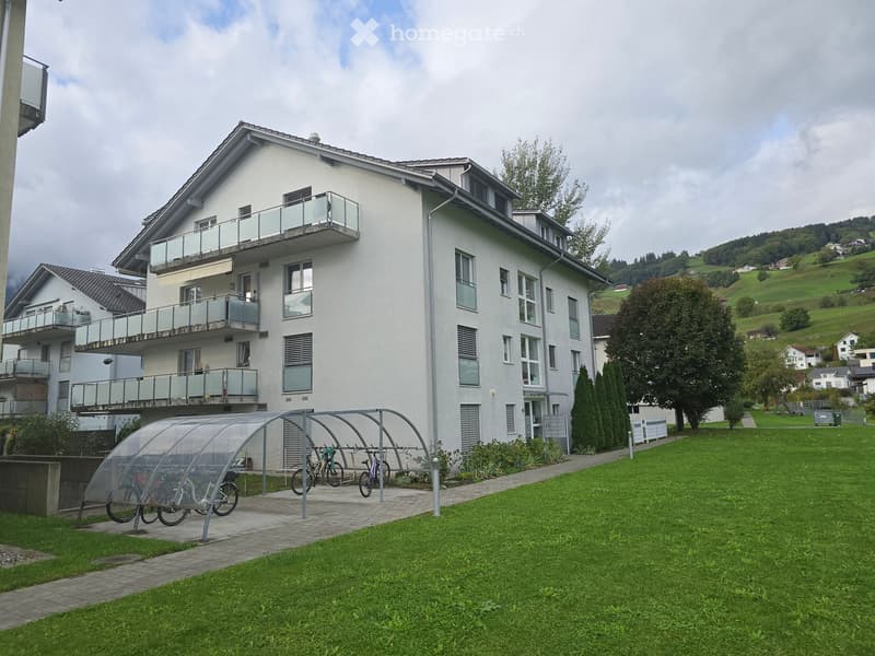 Modern multi-story residential building with balconies, surrounded by a green lawn and a covered bike shelter.
