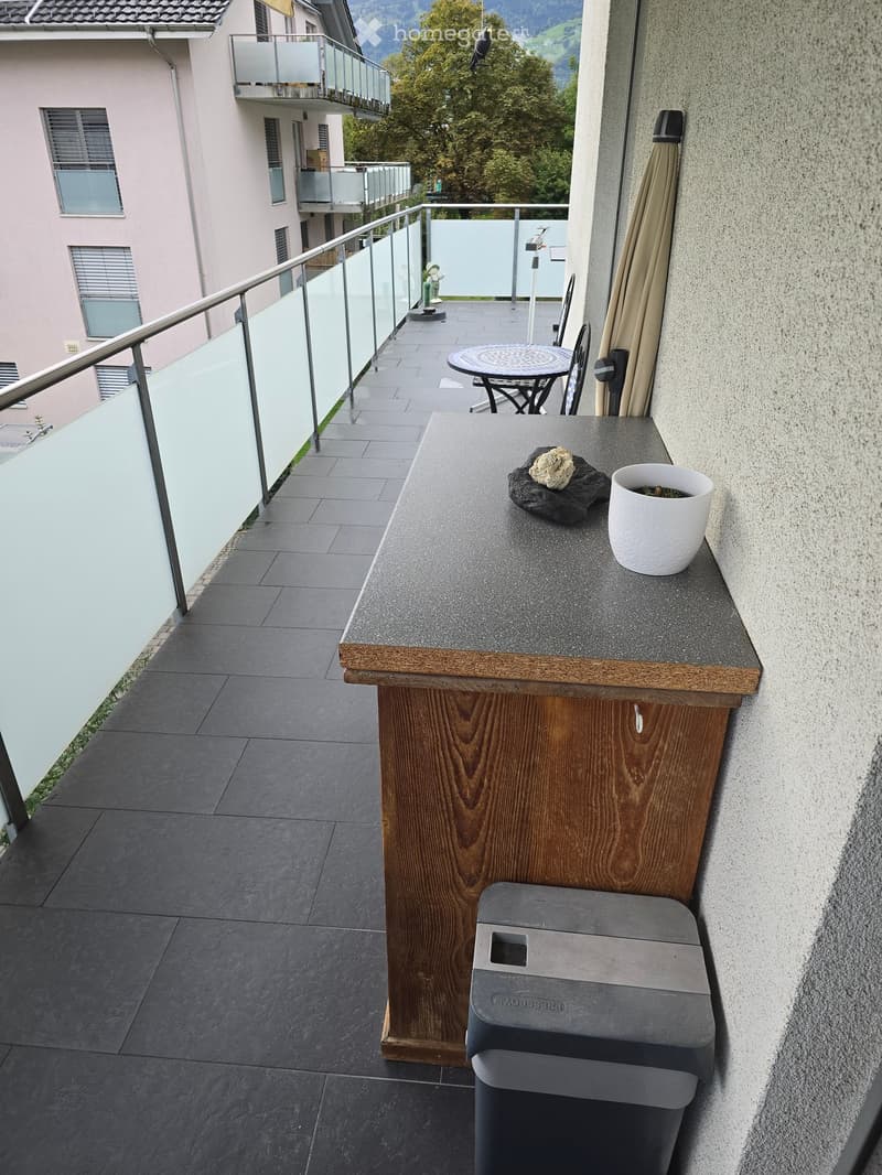 Balcony with grey tiled floor, wooden railing, small table and chair, grey stool, and a white pot on the ledge.