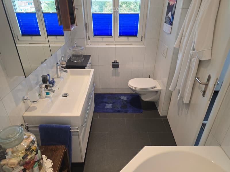 Modern bathroom with a white sink, toilet, bathtub, and mirror. Blue towels are placed on the floor and a small wooden table holds various toiletries and a jar.