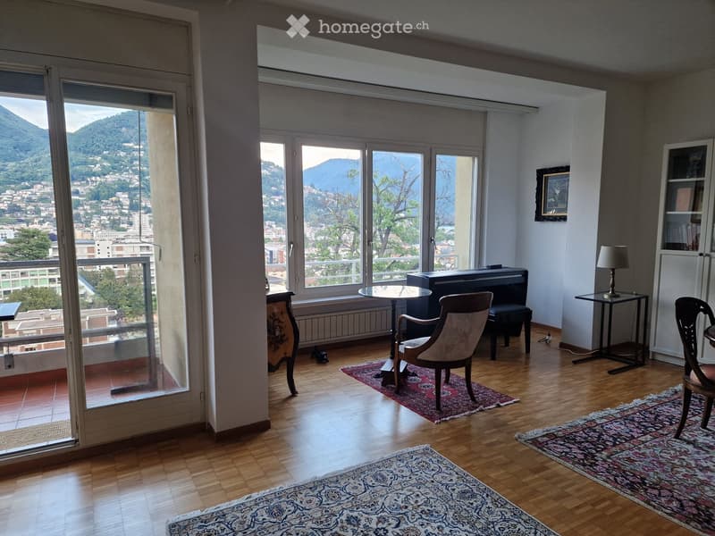 Bright living room with wooden flooring, large windows offering a scenic mountain view, and several pieces of furniture including a chair, table, stool, cabinet, and lamp.