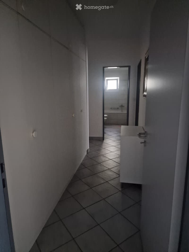 A narrow hallway with tiled flooring and white walls, leading to a small bathroom with a window, toilet, and bathtub.