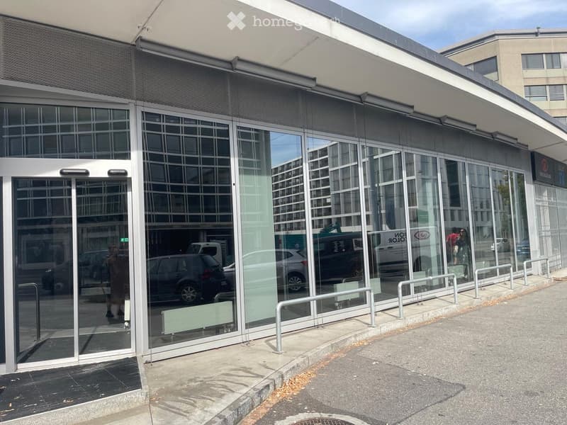 Modern building entrance with large glass doors and reflective windows, featuring a covered walkway and metal railings.