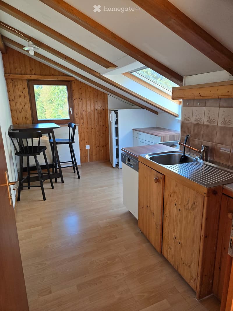 Bright kitchen with wooden cabinets, a sink, refrigerator, and a small dining area with a table and chairs. The room features wooden floors and large windows letting in natural light.