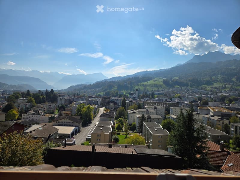 Panoramic view of a town with buildings, trees, and distant mountains under a clear blue sky.