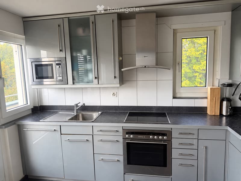 Modern kitchen with grey cabinets, stainless steel appliances, and a black countertop. Features include a microwave, oven, sink, faucet, blender, and cutting board.