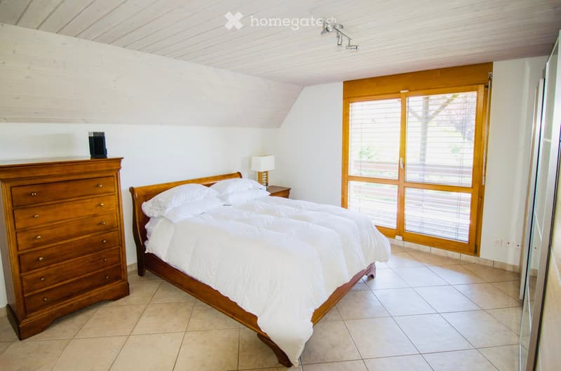 Cozy bedroom with a wooden bed, white bedding, a wooden chest of drawers, a nightstand with a lamp, and an alarm clock on top of the dresser. Large wooden-framed windows with blinds offer natural light.