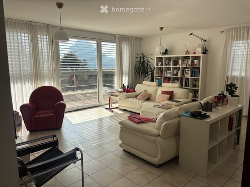 Bright living room with large windows offering a view, cream-colored sofa, red armchair, coffee table, bookshelf, and table lamp.