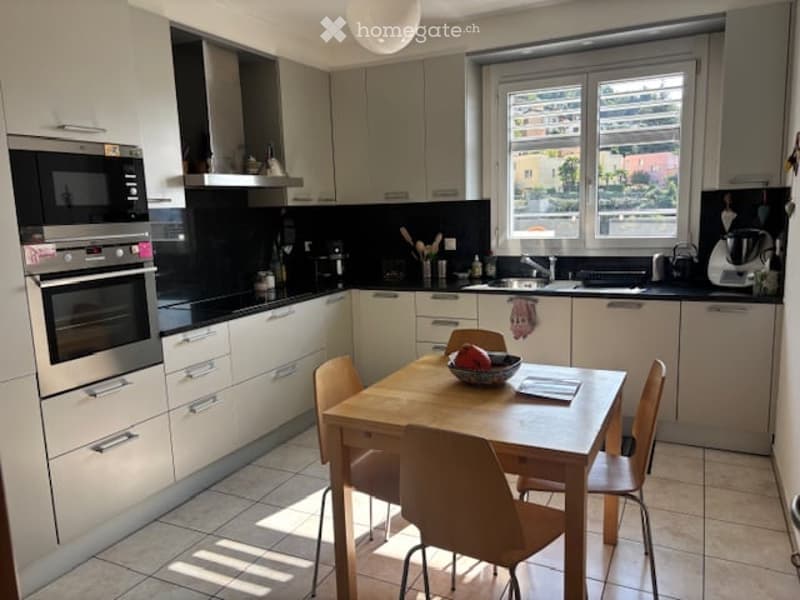 Modern kitchen with white cabinets, black countertops, and a wooden dining table with chairs. Appliances include an oven, microwave, and blender, with a window offering a view of the outside.