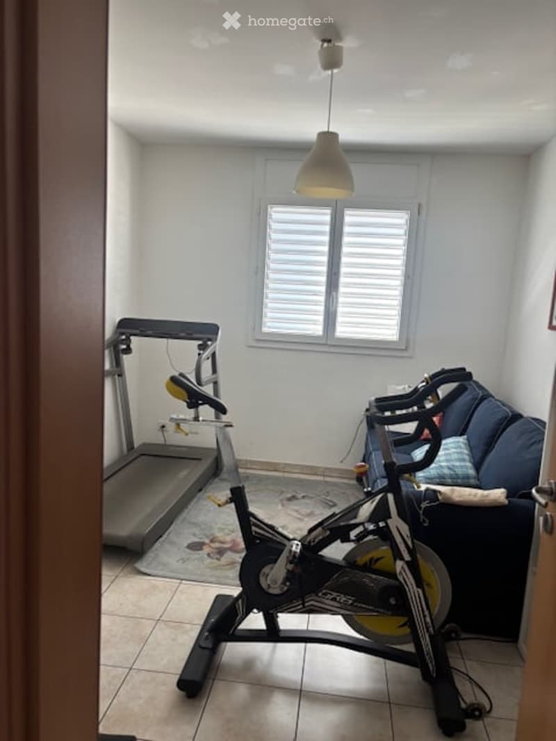 A small, well-lit room with a tiled floor, white walls, and a window with shutters. It contains a sofa with a pillow, an exercise bike, and a treadmill.