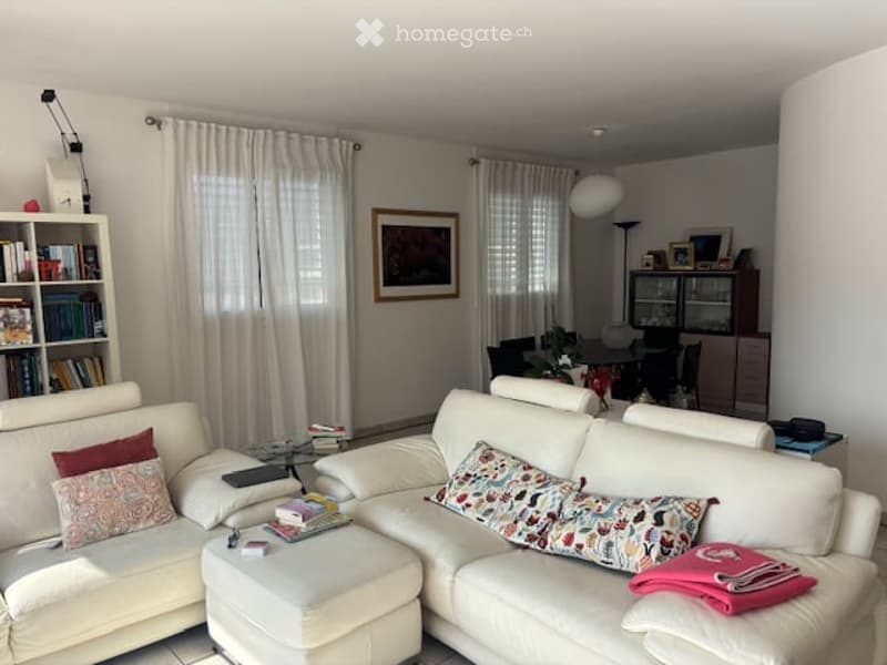 Bright living room with white leather sofa, ottoman, bookshelf, glass coffee table, and dining area with chairs and table.